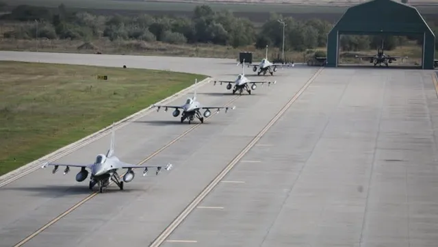 Trei avioane de vânătoare gri taxiază pe o pistă de beton lângă o zonă cu iarbă și un hangar deschis.