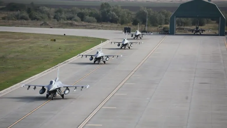 Trei avioane de vânătoare gri taxiază pe o pistă de beton lângă o zonă cu iarbă și un hangar deschis.