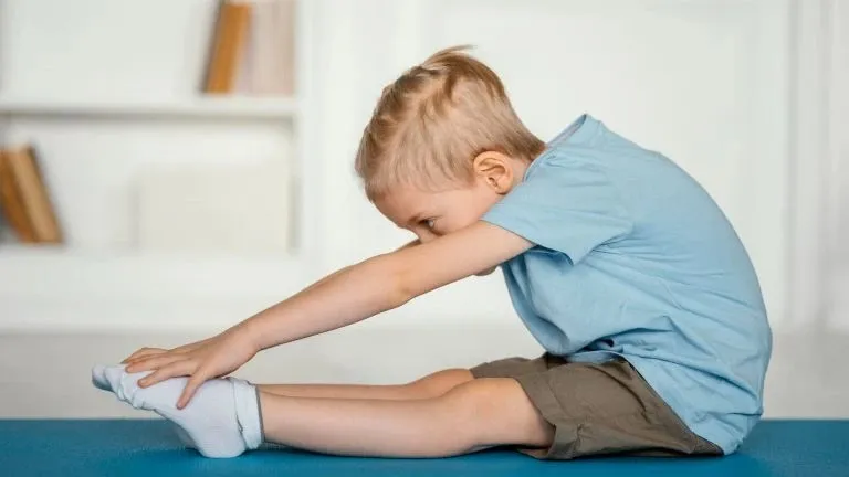 Un copil cu părul blond, purtând un tricou albastru și pantaloni scurți maro, stă pe o suprafață albastră întinzându-se spre picioarele sale.