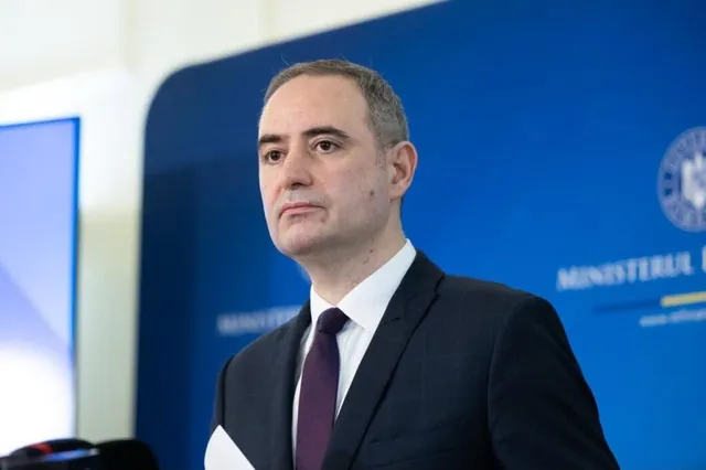 Un bărbat cu părul scurt, purtând sacou închis la culoare, vestă și cravată purpurie, stă în fața unui fundal albastru cu inscripții albe.