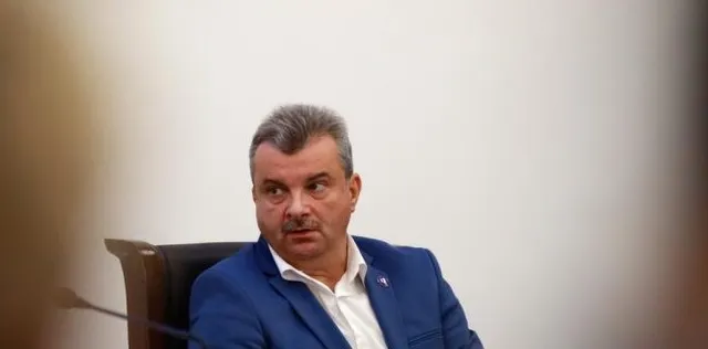 Un bărbat cu părul scurt și mustață, purtând un sacou albastru și cămașă albă, stă pe un scaun de piele maro în fața unui perete alb.
