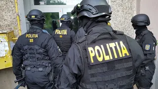Patru polițiști în uniformă neagră cu inscripcții galbene sunt poziționați cu spatele, purtând echipament tactic și căști de protecție.
