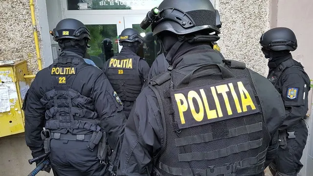 Patru polițiști în uniformă neagră cu inscripcții galbene sunt poziționați cu spatele, purtând echipament tactic și căști de protecție.