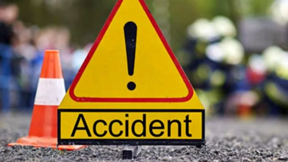 Un panou triunghiular galben cu semnul exclamării și textul "Accident" în prim-plan, alături de un con de trafic portocaliu și alb, pe un fundal neclar.