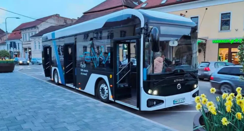 Un autobuz electric alb și negru cu ferestre mari stă oprit pe un drum pavat lângă clădiri și flori galbene.