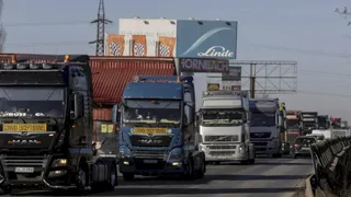 Mai multe camioane de marfă sunt parate pe o bandă de rulare sub un panou publicitar mare sub un cer senin.