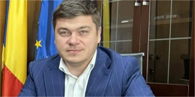 Un bărbat cu părul scurt și deschis la culoare poartă o cămașă albă și un sacou albastru cu dungi subțiri, stând în fața unor steaguri.