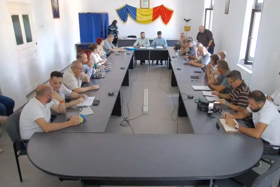 Consilierii locali și invitații sunt așezați la mese în formă de U, în timp ce o cameramană filmează ședința.