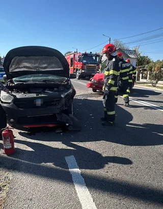 Un autoturism negru cu capota deschisă stă pe un drum asfaltat lângă un stingător, în timp ce pompieri în uniforme de lucru se apropie de un vehicul roșu.