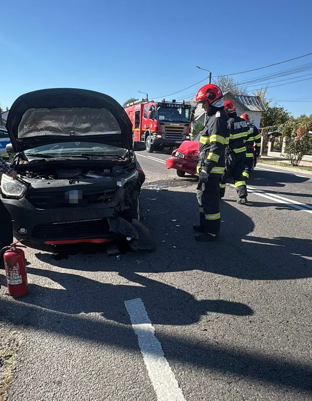 Un autoturism negru cu capota deschisă stă pe un drum asfaltat lângă un stingător, în timp ce pompieri în uniforme de lucru se apropie de un vehicul roșu.