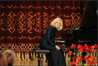 O femeie cu părul blond și rochie neagră stă pe un scaun lângă un pian negru, în fața unui draperiu roșu cu modele aurii.