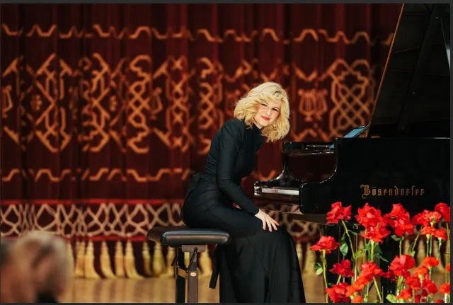 O femeie cu părul blond și rochie neagră stă pe un scaun lângă un pian negru, în fața unui draperiu roșu cu modele aurii.