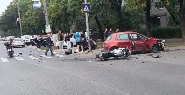 Un autoturism roșu este oprit pe carosabil lângă o motocicletă răsturnată, în timp ce mai mulți oameni stau în apropierea vehiculelor pe stradă.