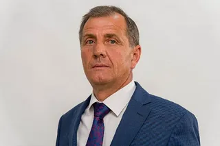 Un bărbat cu părul scurt, purtând sacou albastru, cămașă albă și cravată cu modele colorate, privește spre cameră pe un fundal alb.