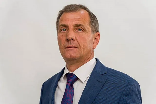 Un bărbat cu părul scurt, purtând sacou albastru, cămașă albă și cravată cu modele colorate, privește spre cameră pe un fundal alb.