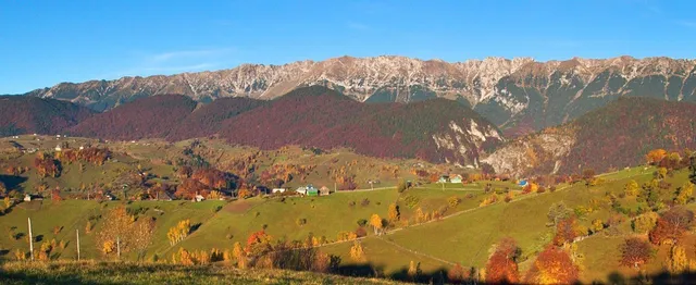 Munți cu creste stâncoase sub un cer albastru deasupra unor dealuri verzi acoperite de vegetație cu frunze colorate și câteva case mici.