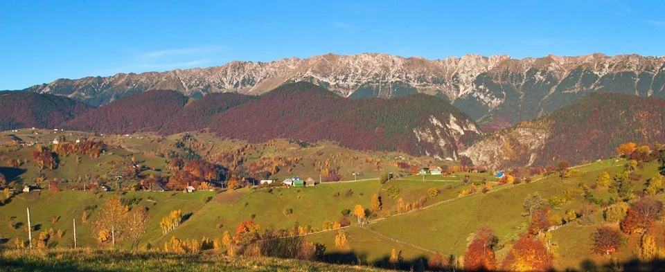 Munți cu creste stâncoase sub un cer albastru deasupra unor dealuri verzi acoperite de vegetație cu frunze colorate și câteva case mici.