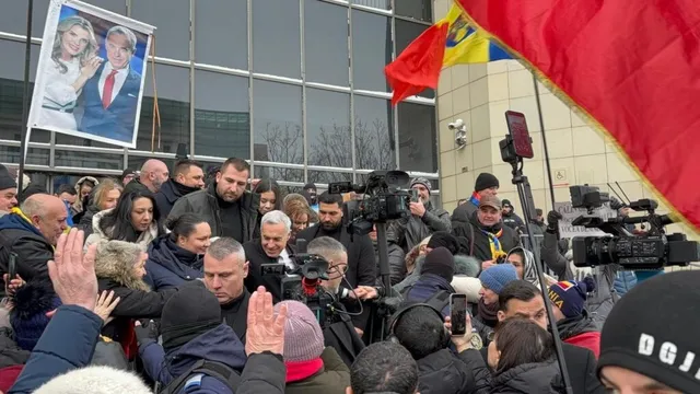 O mulțime de oameni cu haine de iarnă stă în fața unei clădiri cu ferestre mari, sub un poster și un steag roșu.