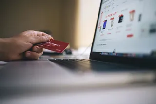 O mână ține un card roșu în fața unui ecran de laptop care afișează un magazin online cu diverse produse.