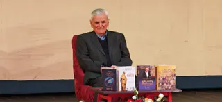 Un bărbat cu părul alb, îmbrăcat într-un sacou închis și un pulover, stă pe un scaun roșu lângă o masă cu patru cărți.