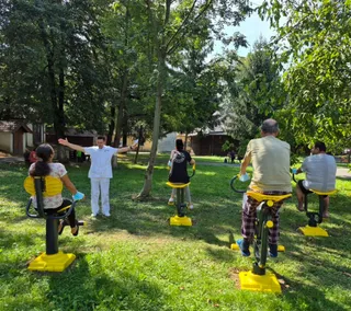 Un grup de persoane participă la o activitate în aer liber, folosind aparate de exerciții galbene pe o peluză verde înconjurată de copaci.