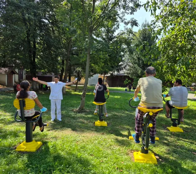 Un grup de persoane participă la o activitate în aer liber, folosind aparate de exerciții galbene pe o peluză verde înconjurată de copaci.