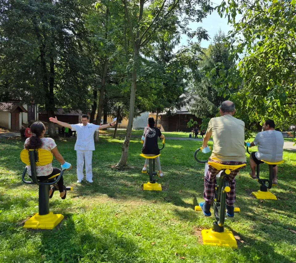 Un grup de persoane participă la o activitate în aer liber, folosind aparate de exerciții galbene pe o peluză verde înconjurată de copaci.