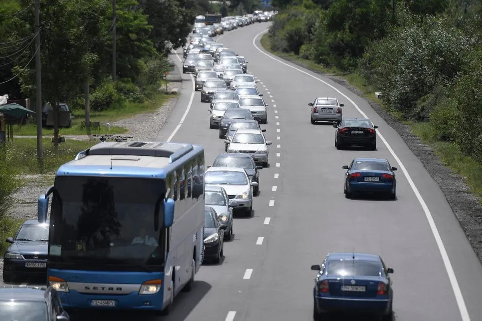 Un autobuz albastru și alb ocupă partea stângă a unui drum asfaltat, în timp ce mai multe mașini circulă pe banda din dreapta și în trafic.