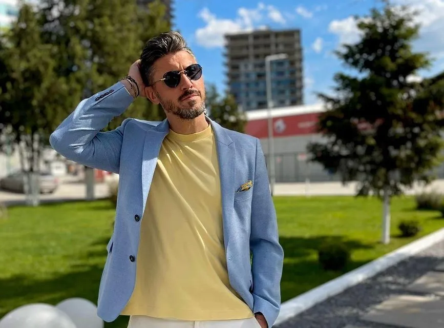 Un bărbat cu ochelari de soare, purtând un sacou albastru și un tricou galben, stă în picioare pe un teren verde cu clădiri în fundal.