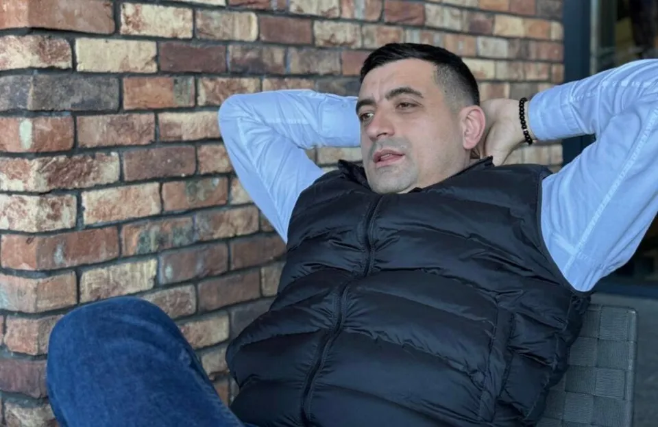 Un bărbat cu părul scurt, purtând o cămașă albastră și o vestă neagră, stă sprijinit de un perete de cărămidă cu mâinile la spate capului.