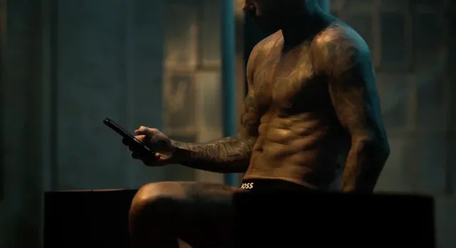 Un bărbat musculos, fără tricou, stă așezat și ține un dispozitiv electronic în mâna stângă.