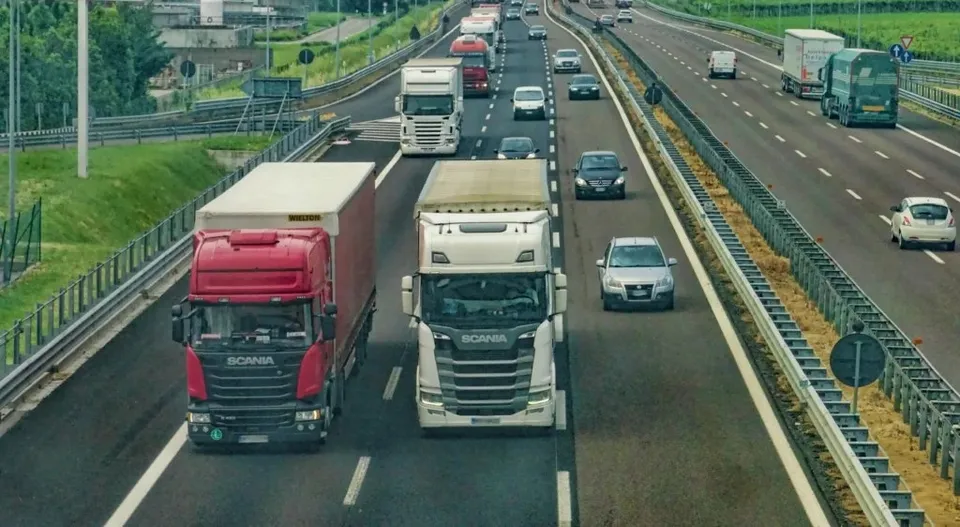 Mai multe camioane și mașini circulă pe o autostradă cu mai multe benzi de circulație, întreagă fiind delimitată de bariere metalice.