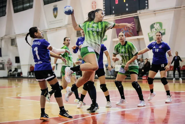 Femei jucând handbal pe un teren de interior, dintre care una sare cu o minge albastră în mână, fiind înconjurată de alte jucătoare.