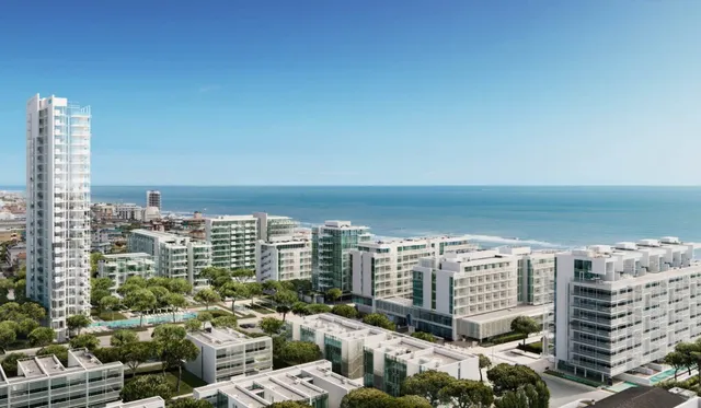 O vedere aeriană a unui oraș de coastă cu clădiri albe moderne, copaci verzi și marea sub un cer senin.