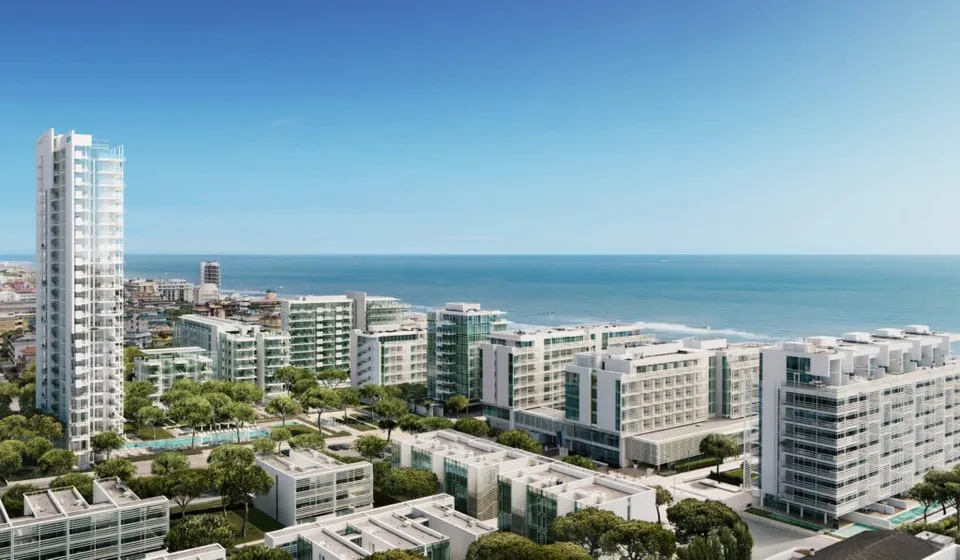 O vedere aeriană a unui oraș de coastă cu clădiri albe moderne, copaci verzi și marea sub un cer senin.