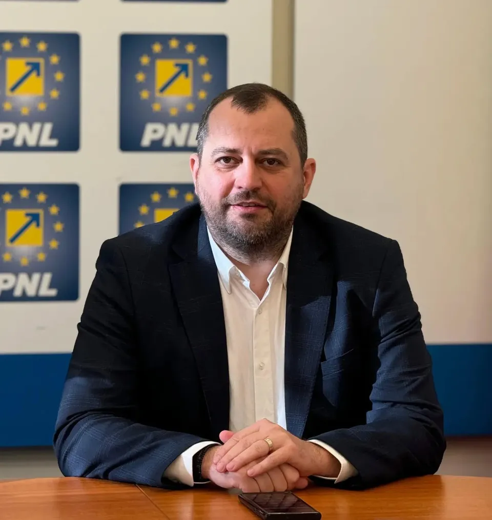 Un bărbat cu barbă, purtând sacou închis și cămașă albă, stă la o masă de lemn în fața unor panouri cu logo-uri albastre și galbene.
