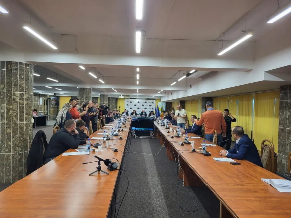 Oamenii stau la mese lungi de lemn într-o sală de ședințe cu lumini albe suspendate și un ecran de monitor în fundal.