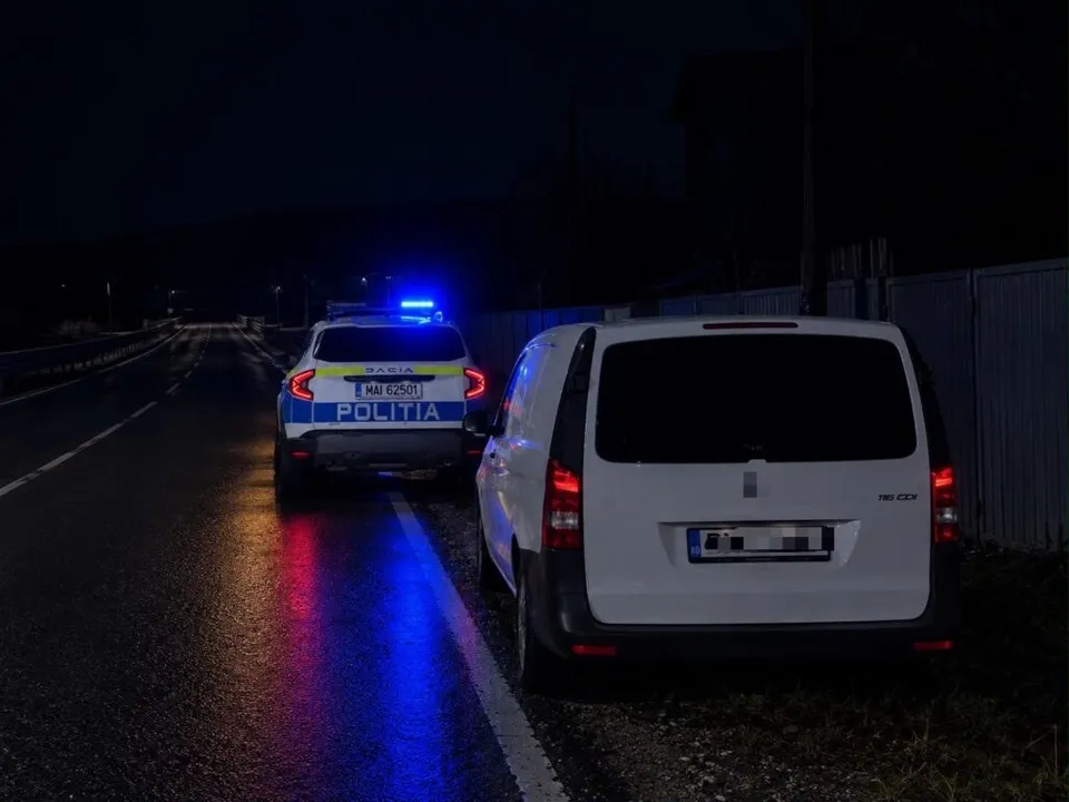 Un vehicul de poliție cu girofaruri albastre aprinse stă oprit pe marginea unui drum asfaltat lângă o mașină albă de tip van.