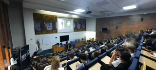 O sală de conferințe mare cu spectatori așezați pe rânduri de scaune albastre în fața unui ecran de proiecție și a unei mese de prezentare.