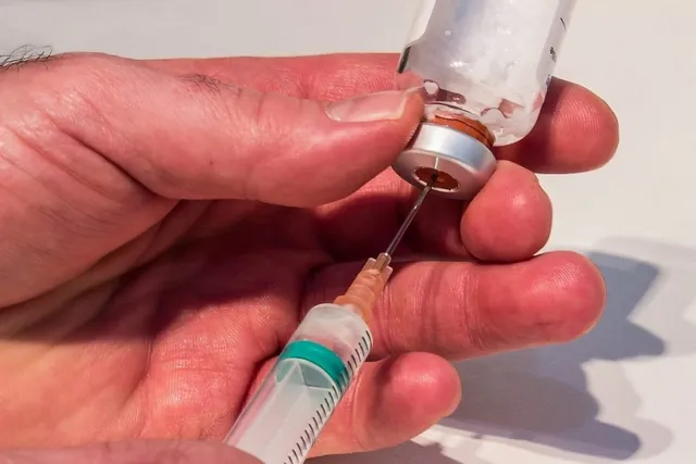 O mână ține un flacon de sticlă transparent cu lichid și introduce un ac de seringă cu marcaje verzi în dopul metalic al acestuia.