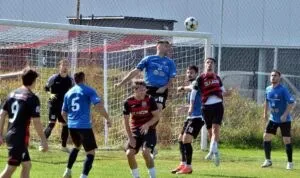 Cei opt jucători de fotbal de pe teren sunt în poziții de joc, unii privind spre un balon aflat în aer în apropierea porții.