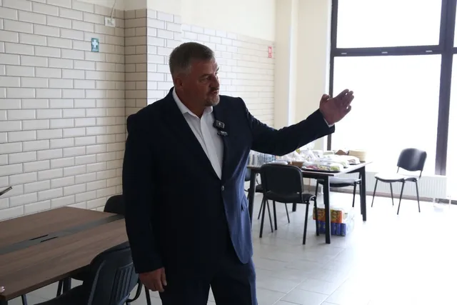 Un bărbat cu părul scurt și costum închis stă în picioare într-o încăpere cu pereți din cărămidă albă, întinzând mâna dreaptă spre o masă.
