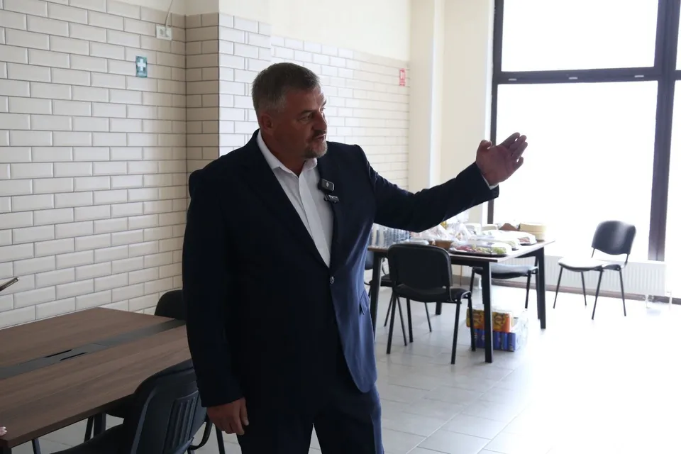 Un bărbat cu părul scurt și costum închis stă în picioare într-o încăpere cu pereți din cărămidă albă, întinzând mâna dreaptă spre o masă.