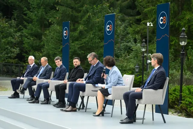 Oamenii stau așezați într-un rând pe scaune gri pe o platformă înaltă, în fața unor panouri albastre cu inscripții circulare albe și portocalii.