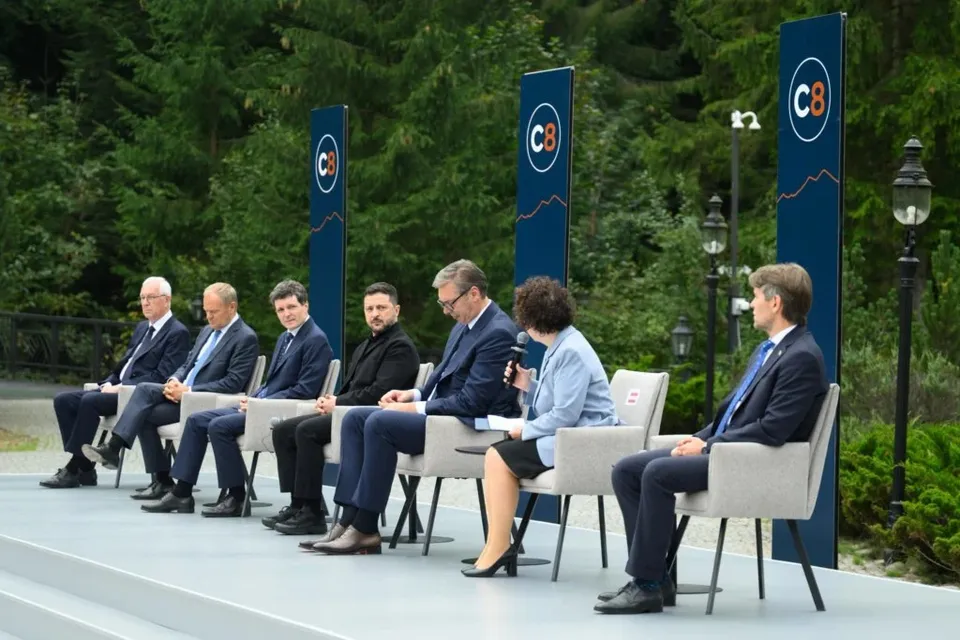 Oamenii stau așezați într-un rând pe scaune gri pe o platformă înaltă, în fața unor panouri albastre cu inscripții circulare albe și portocalii.