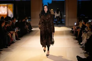 O femeie merge pe o pistă de catwalk purtând o rochie lungă cu imprimeu leopard, pantofi negri și ținând o geantă mică în mână.