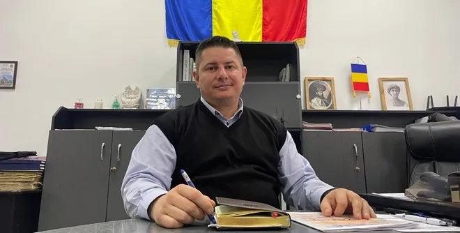 Un bărbat cu cămașă albastră și vestă neagră stă la o masă cu documente și un pix în mână, în fața unor dulapuri gri și un steag.