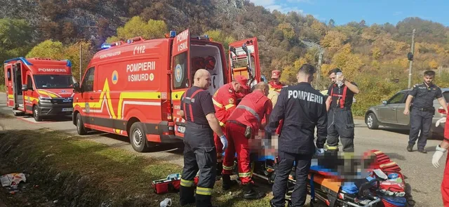 Echipe de pompieri în uniforme roșii și negre lucrează lângă un vehicul de urgență roșu pe marginea unui drum asfaltat.