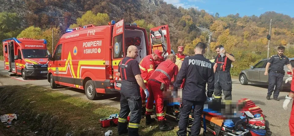 Echipe de pompieri în uniforme roșii și negre lucrează lângă un vehicul de urgență roșu pe marginea unui drum asfaltat.