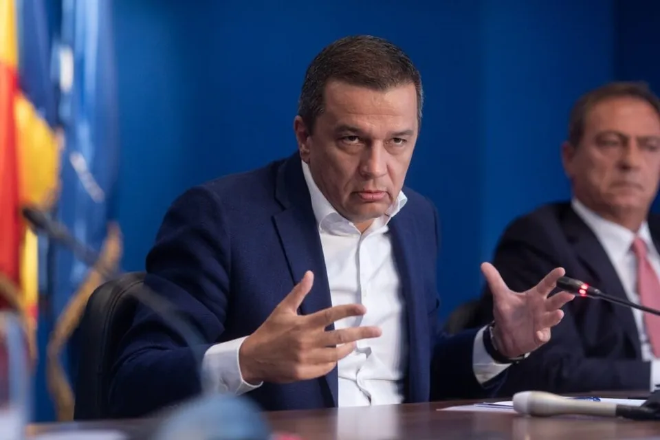 Un bărbat într-un costum albastru și cămașă albă stă la o masă și face gesturi cu ambele mâini în fața unor microfoane.